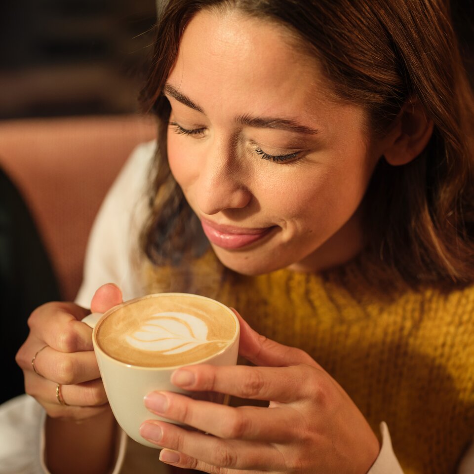 Junge Frau genießt einen Milchkaffee mit Herz-Latte-Kunstwerk und trägt einen gemütlichen Strickpullover in einem Café.