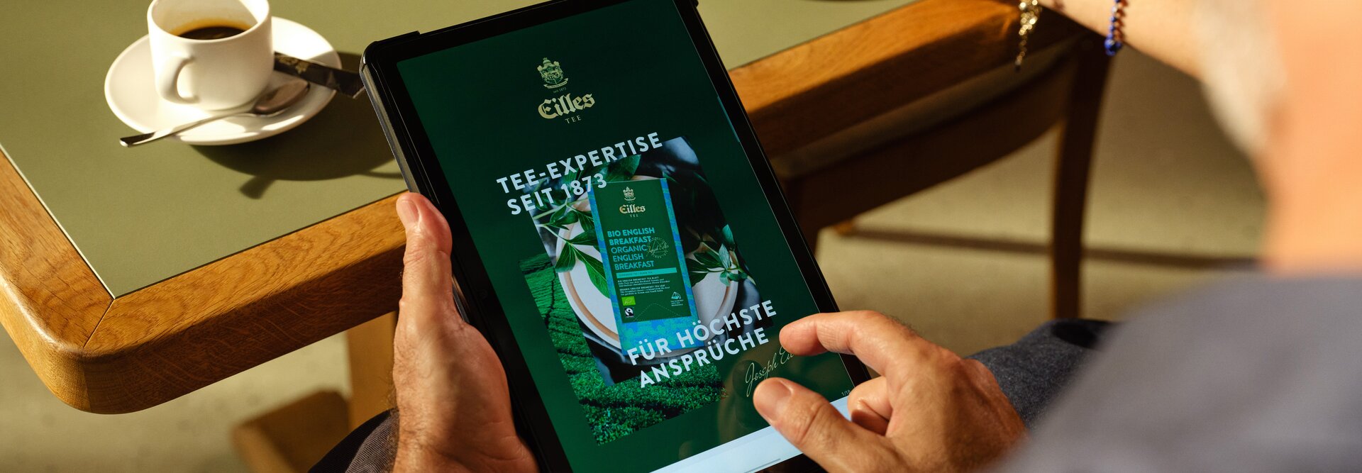 Eine Person genießt Tee und Kaffee, während sie auf einem Tablet an einem Tisch in der Eilles Tea Expertise stöbert.