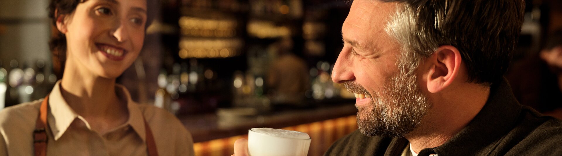 Ein lächelnder Barista serviert einem glücklichen Kunden einen Milchkaffee in einer warmen, einladenden Caféumgebung. Perfekt für Kaffee-Liebhaber.