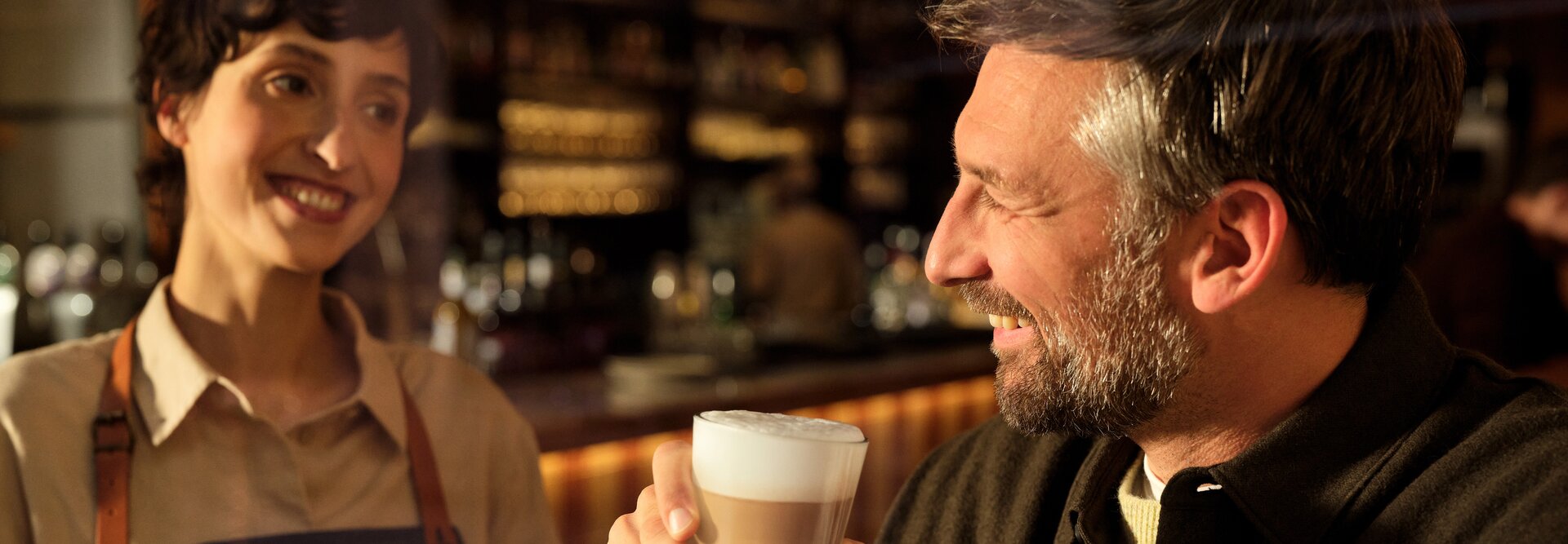 Ein lächelnder Barista serviert einem glücklichen Kunden einen Milchkaffee in einer warmen, einladenden Caféumgebung. Perfekt für Kaffee-Liebhaber.