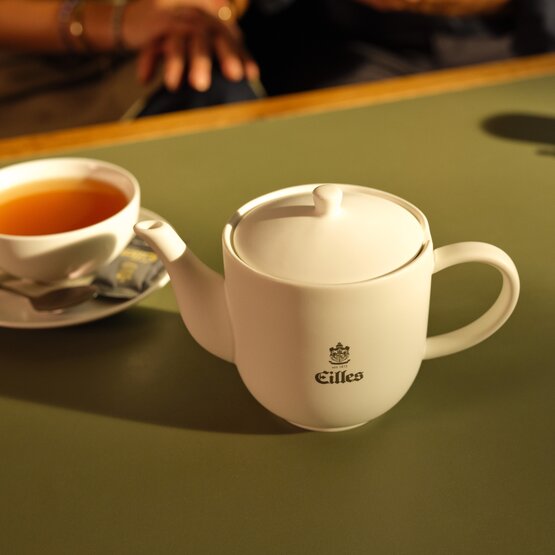 Witte Eilles theepot naast een kopje gezette thee op een groene tafel, perfect voor theemomenten in de namiddag.