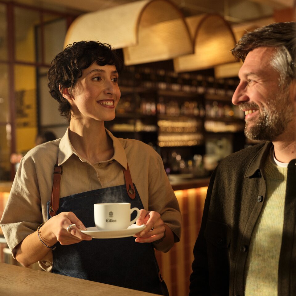 Ein Barista serviert einem lächelnden Kunden in einem lebhaften Café eine dampfende Tasse Eilles-Kaffee.