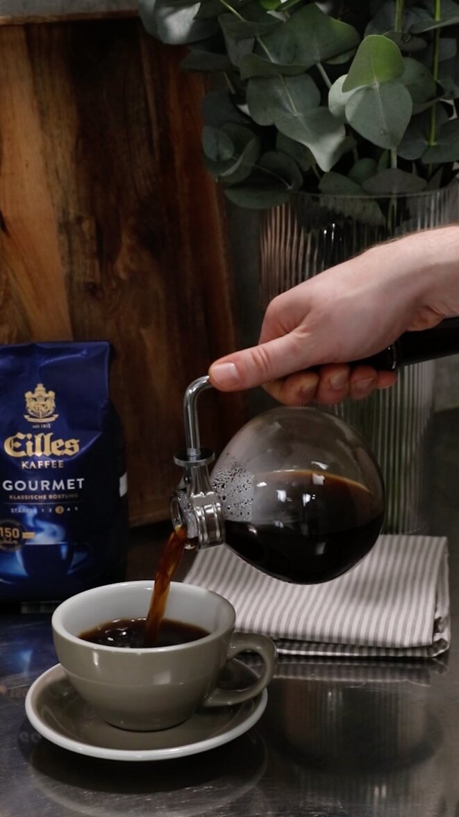 Einfüllen von frisch gebrühtem Kaffee aus einem Siphon in eine Tasse, umgeben von Eilles-Kaffee und Eukalyptusblättern.