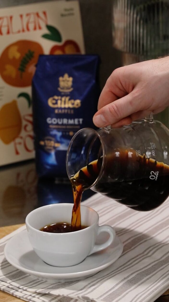 Pouring freshly brewed coffee from a glass jug into a white cup on a striped cloth, with gourmet coffee bag in background.