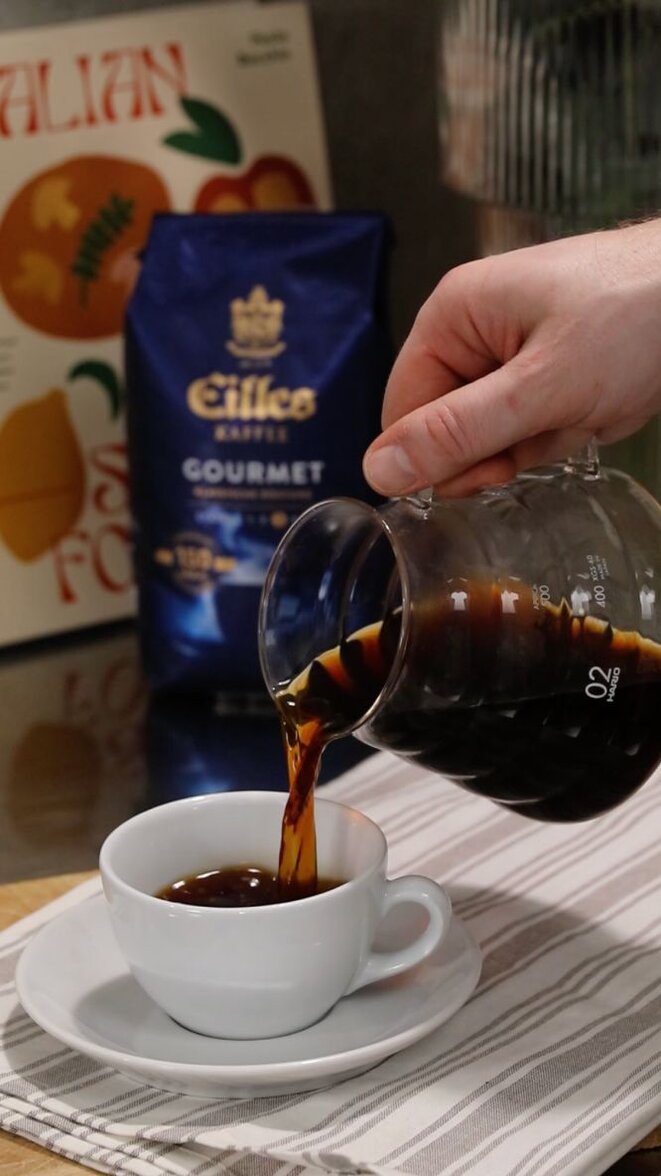 Einschenken von frisch gebrühtem Kaffee aus einer Glaskanne in eine weiße Tasse auf einem gestreiften Tuch, mit einer Gourmet-Kaffeetüte im Hintergrund.