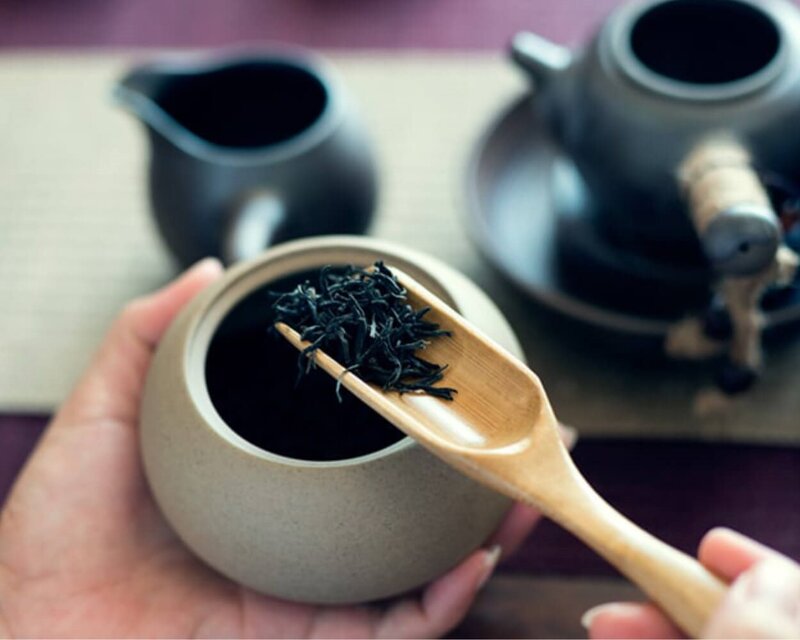 Loser schwarzer Tee wird mit einem Bambuslöffel aus einem dekorativen Behälter abgemessen. Im Hintergrund ist eine Teekanne zu sehen.