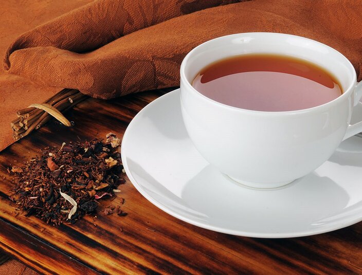 Čaj Rooibos v bílém šálku vedle sypaných lístků na dřevěném podnosu s texturovaným hadříkem. Ideální pro příjemné odpoledne.