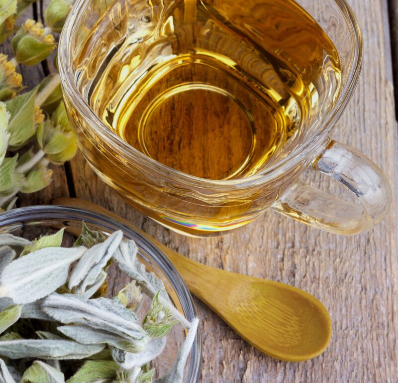 Kräutertee, serviert in einer Glastasse, begleitet von getrockneten Kräutern und einem Holzlöffel auf einer rustikalen Holzunterlage.