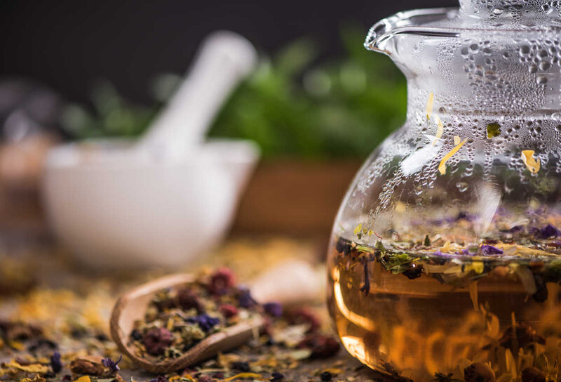 Eine gläserne Teekanne, gefüllt mit duftendem Früchtetee, umgeben von Kräutern und Trockenblumen auf einem Holztisch.