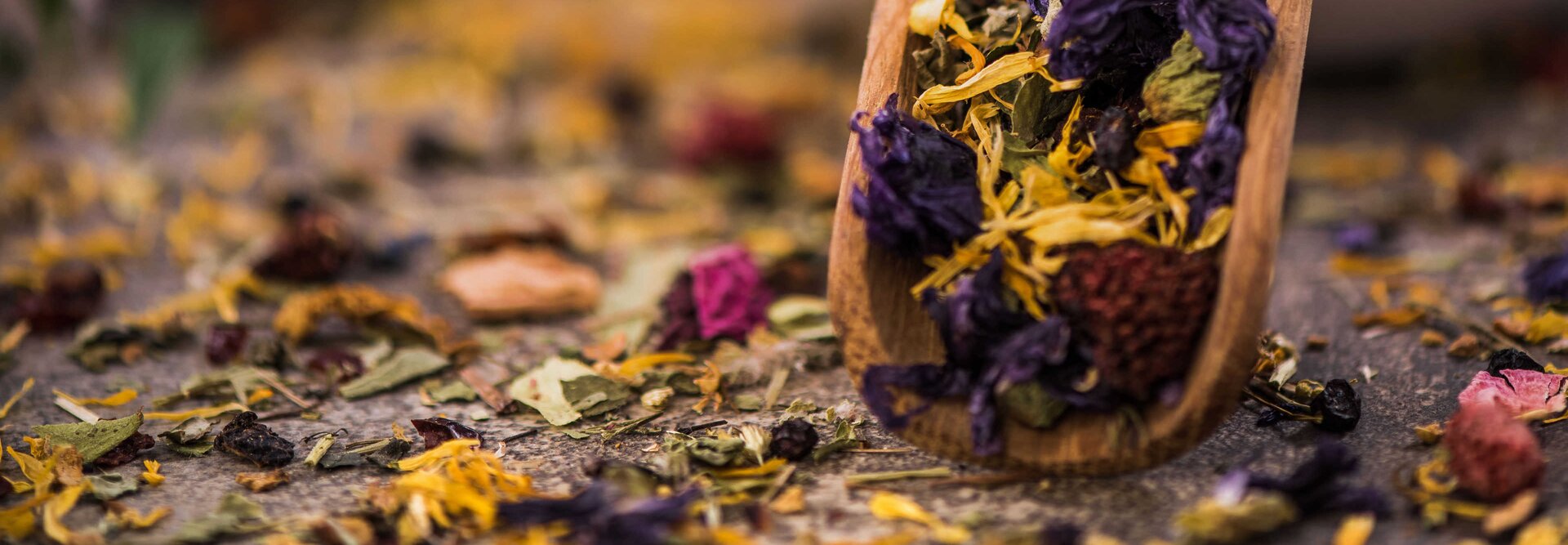 Eine farbenfrohe Kräuterteemischung, die aus einer hölzernen Schöpfkelle fließt und getrocknete Blüten und Blätter auf einer rustikalen Oberfläche präsentiert.
