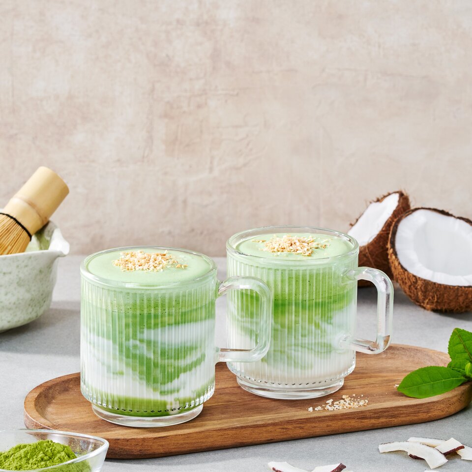 Köstlicher heißer Kokosnuss-Matcha-Latte, serviert in Glaskrügen, garniert mit Kokosnussflocken und Grünteepulver.