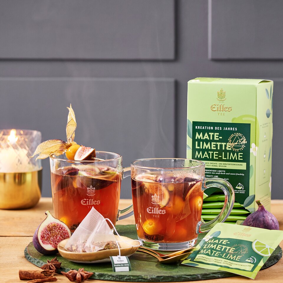 Warmer Mate-Limette-Tee, garniert mit Feigen, serviert in Glaskrügen zusammen mit der Eilles-Tee-Verpackung auf einem Holztisch.