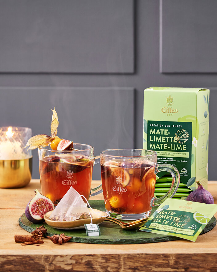 Warmer Mate-Limette-Tee, garniert mit Feigen, serviert in Glaskrügen zusammen mit der Eilles-Tee-Verpackung auf einem Holztisch.
