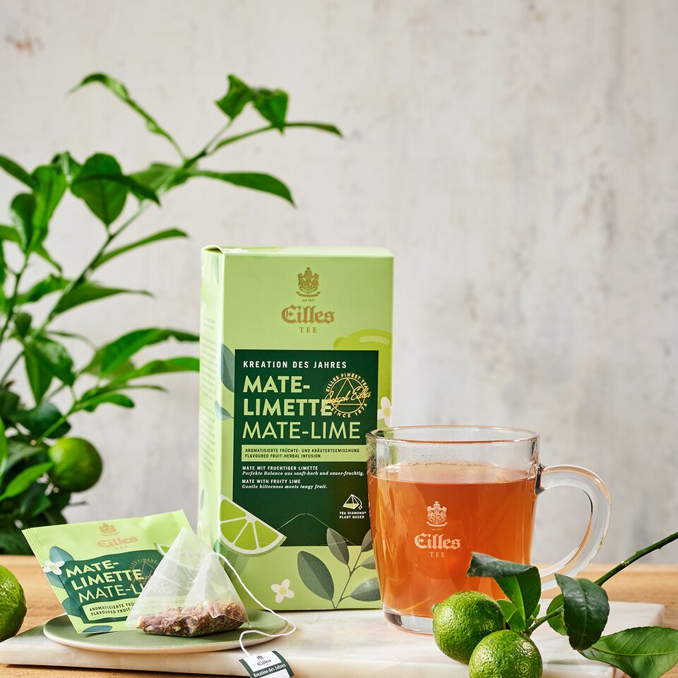 Erfrischende Eilles-Tee-Packung mit Mate-Limette, mit aufgebrühtem Tee und frischen Limetten auf einer Holzfläche, umgeben von Grün.