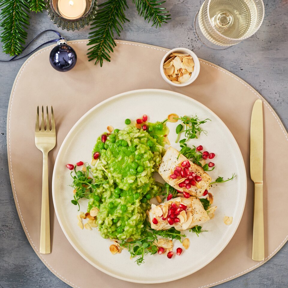 Grünes Erbsenrisotto mit Kabeljau, garniert mit Granatapfel und Mandeln, angerichtet auf einem stilvollen Tisch mit festlicher Dekoration.