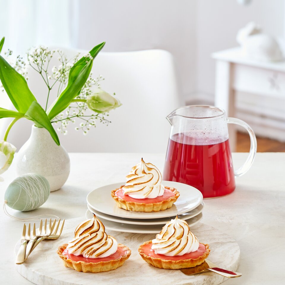 Wunderschöne Zitronentörtchen mit geröstetem Baiser, serviert mit einem Krug Beerengetränk und dekorativen Blumen auf einem Tisch.