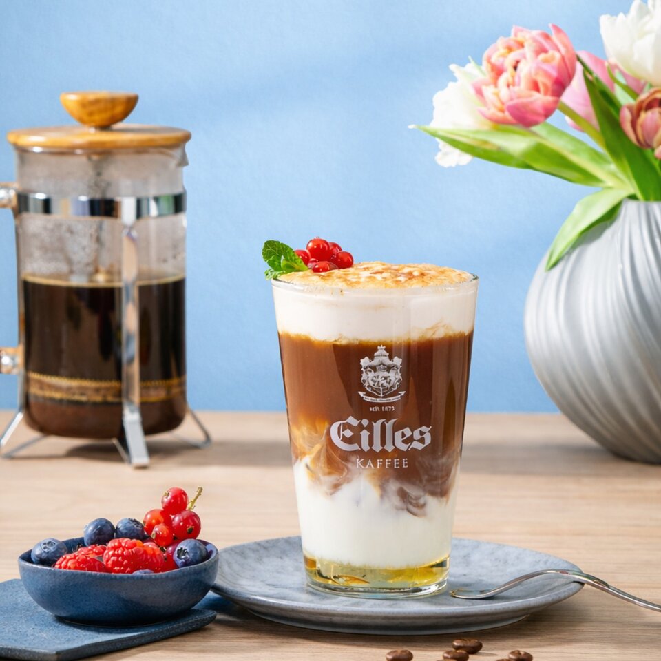 Eiskaffee mit Crème brûlée, garniert mit Beeren und Minze, serviert mit einer französischen Presse und einem Blumenarrangement.