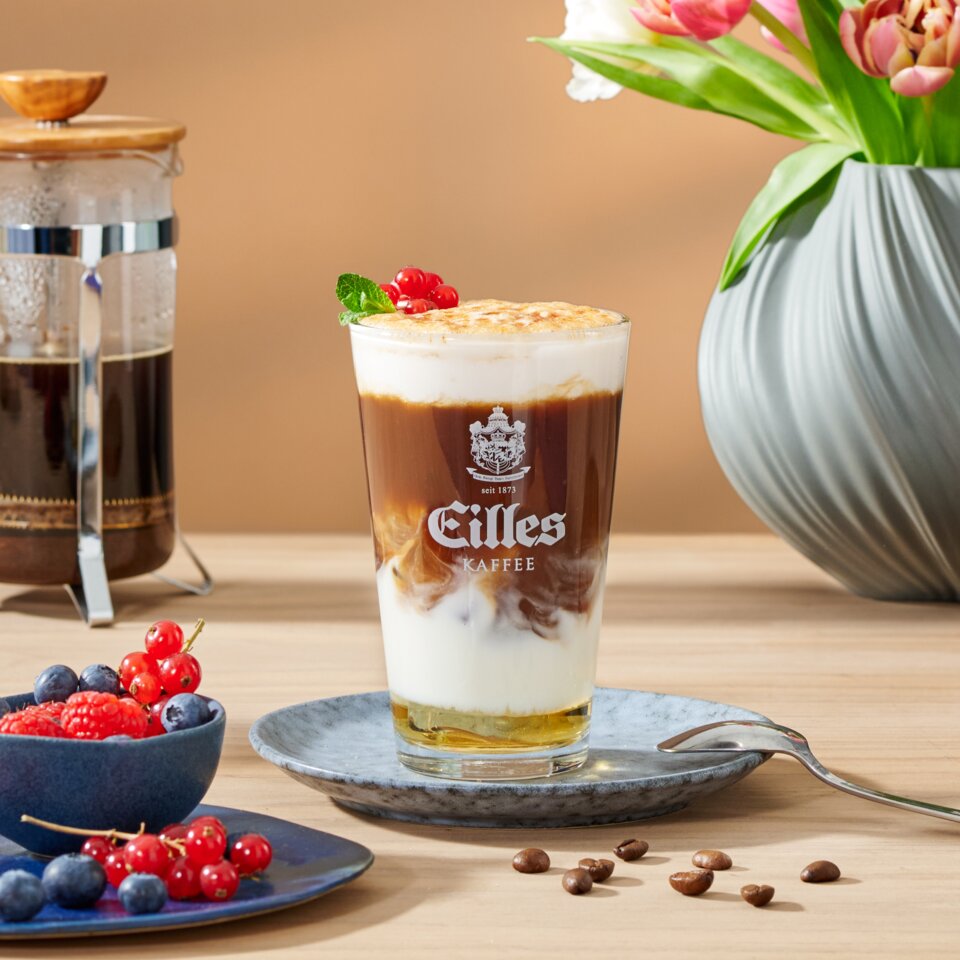 Delicious crème brûlée iced latte with fresh berries and flowers, next to a French press and coffee beans.