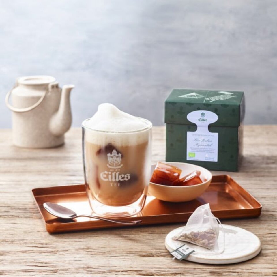 Köstliche Tee- und Kaffeemischung von Eilles, serviert in einem Glas, begleitet von Karamellwürfeln und einer Teebox auf einem Holztablett.