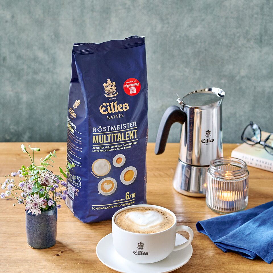 Eilles Röstmeister Multitalent Kaffee, Cappuccino, Kanne und Blumen auf einem Holztisch in gemütlicher Umgebung.
