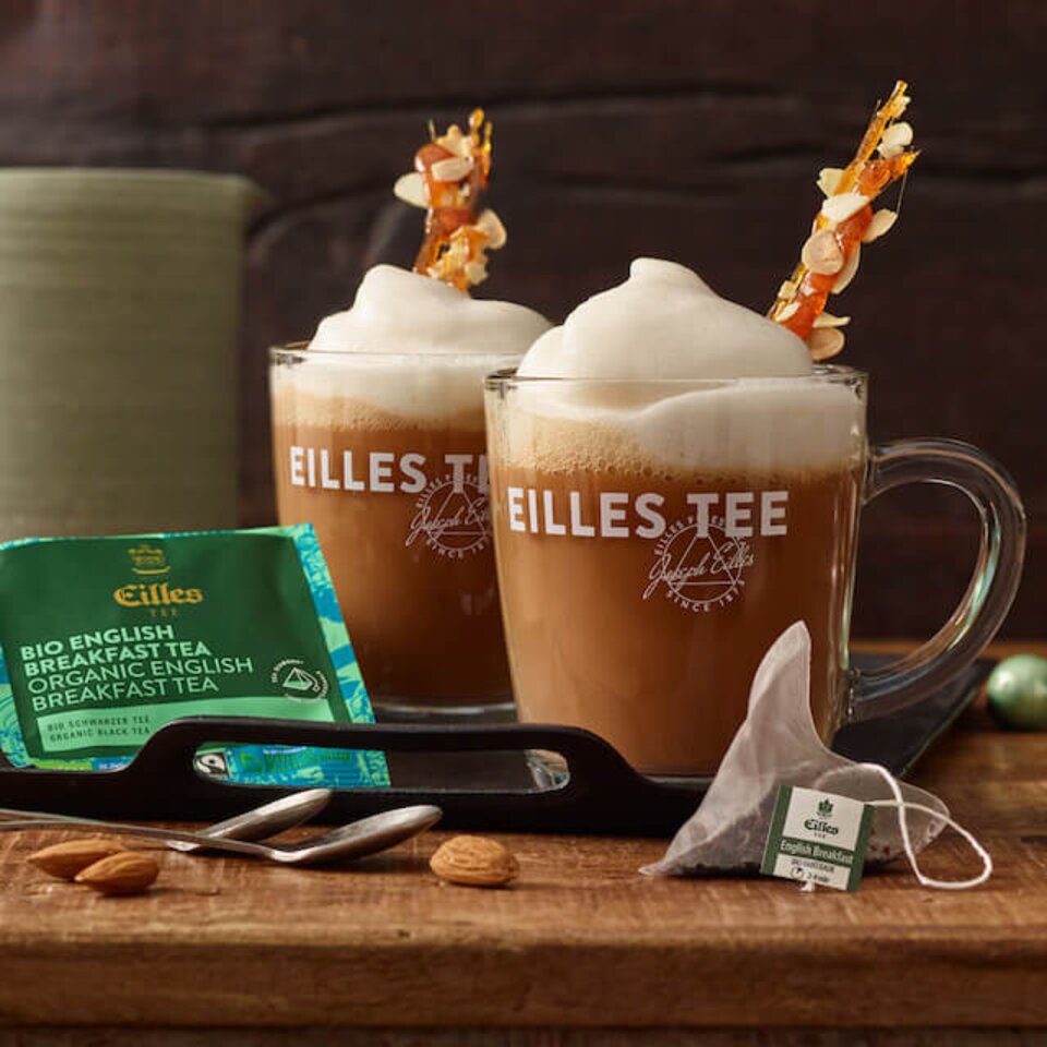 Englischer Bio-Frühstückstee, serviert in schaumigen Tassen, begleitet von Garnierungen und Teebeuteln auf einer Holzunterlage.
