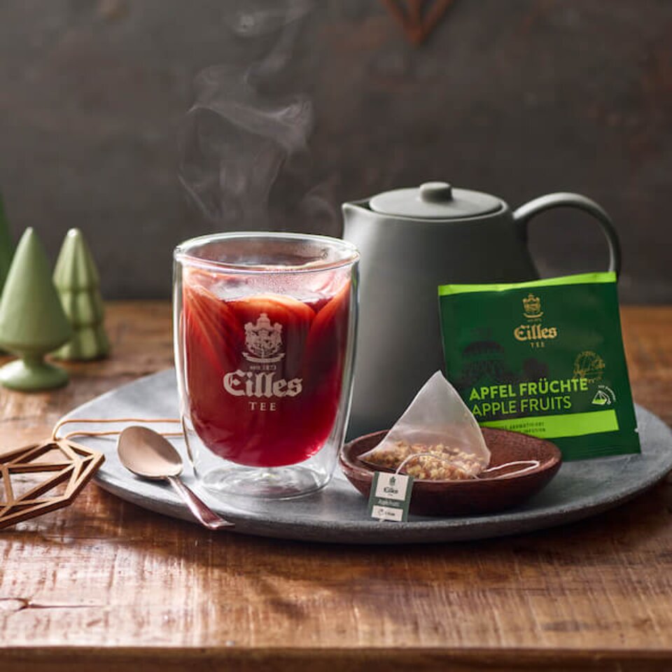 Eine dampfende Tasse Apfel-Früchtetee neben einer Teekanne und losem Tee mit einem Holzlöffel auf einem rustikalen Tablett.