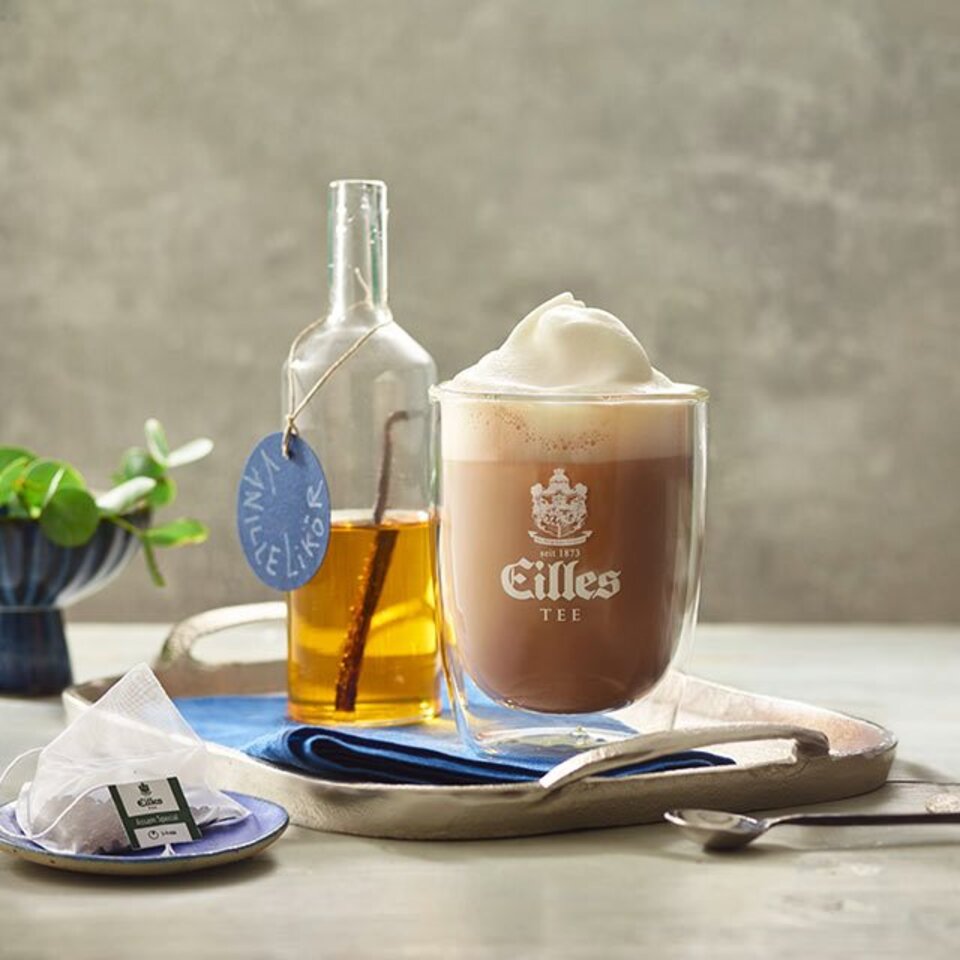 Cremiger Assam-Hafermilchpunsch, serviert in einem Glas mit Eilles Tee, zusammen mit einer Flasche Sirup und einem Teebeutel.