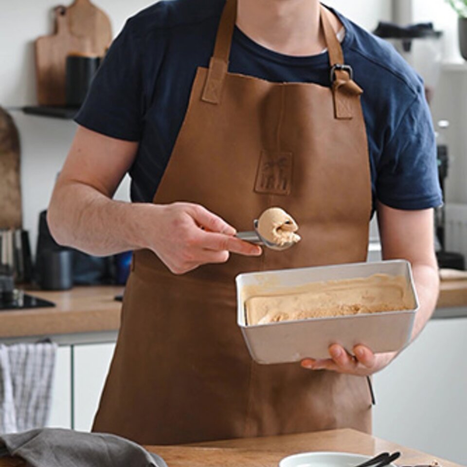 Ein Mann in einer braunen Schürze löffelt Kaffee-Eis in einer hellen Küche, umgeben von Eiswaffeln und Toppings.
