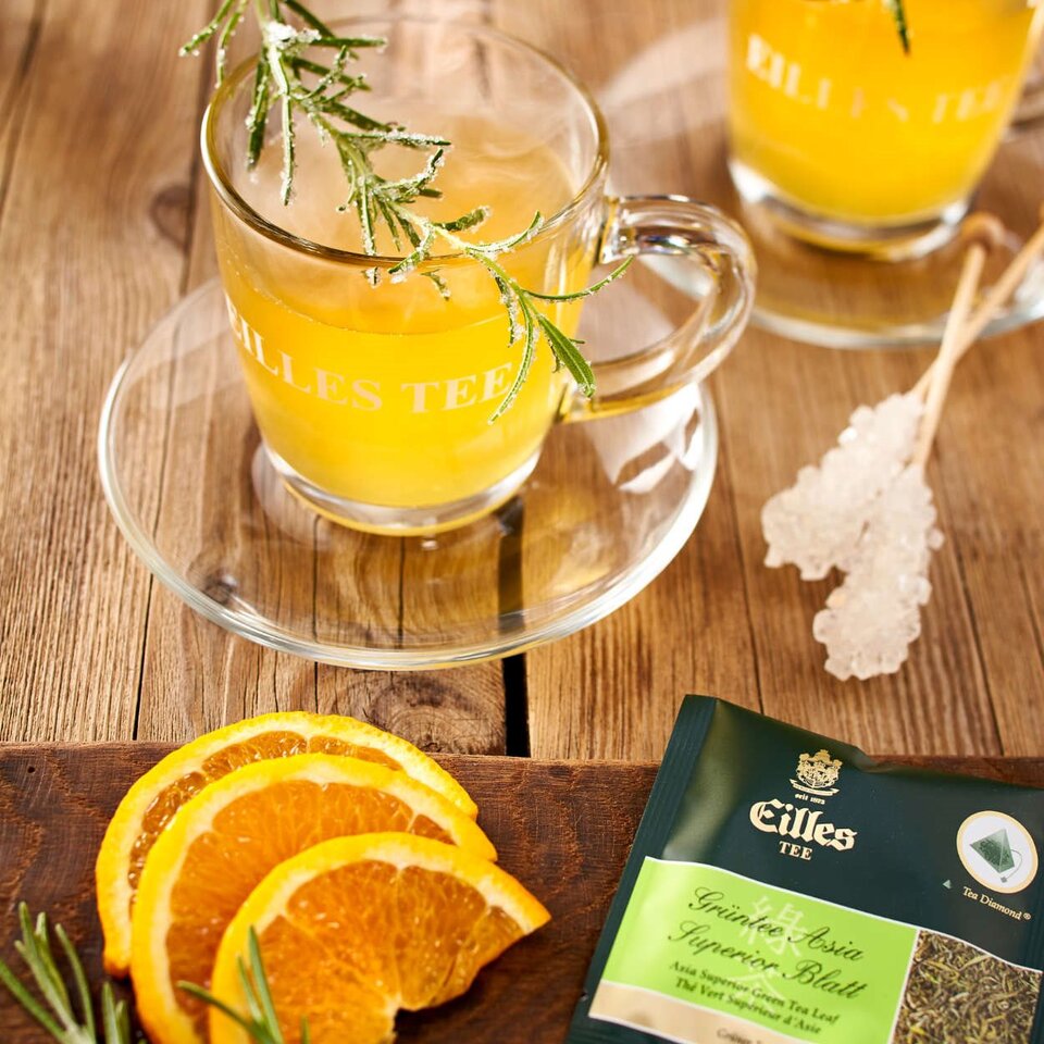 Dampfender Kräutertee mit Rosmarin und Orangenscheiben, gepaart mit einer Eilles-Tee-Verpackung auf einem rustikalen Holztisch.