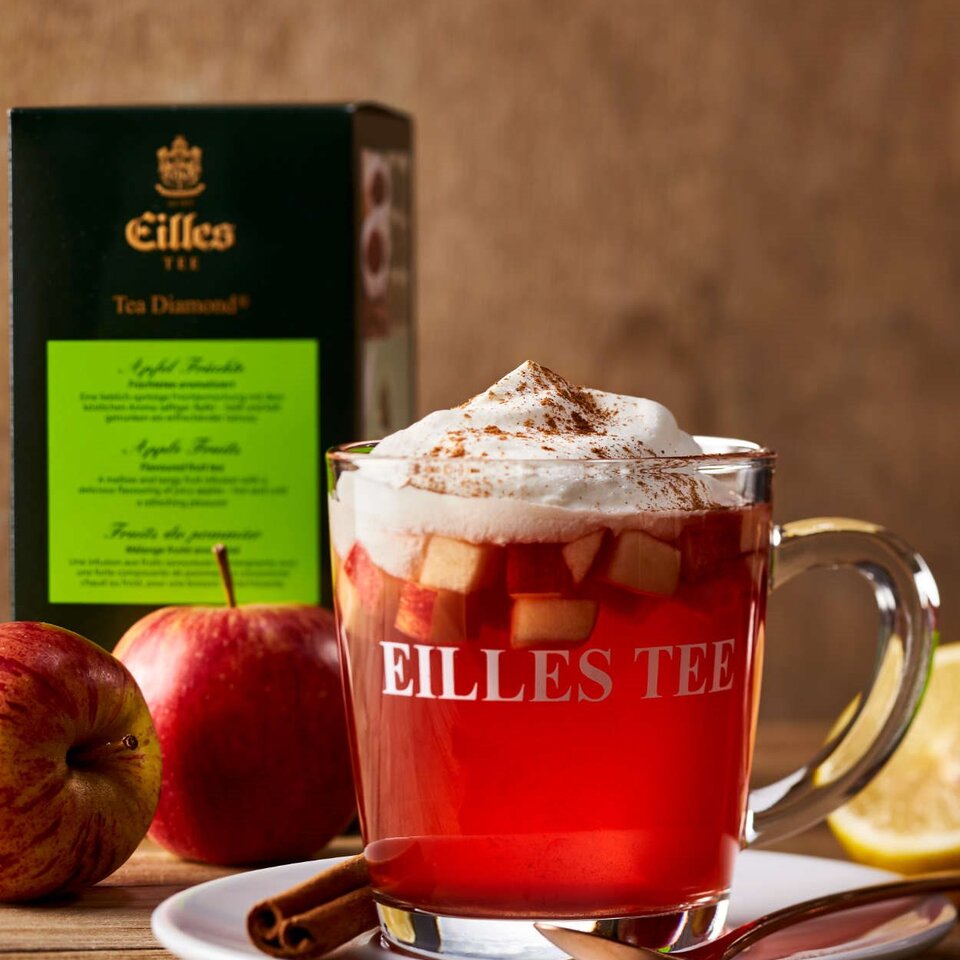 Warmer Apfeltee mit Sahne und Gewürzen, umgeben von frischen Äpfeln und Eilles Teebox. Perfekt für gemütliche Wintergetränke.