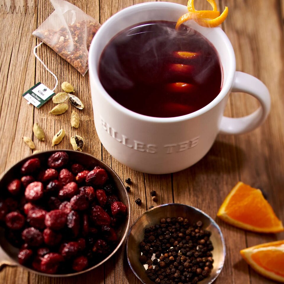 Eine dampfende Tasse mit Cranberry-Orangen-Punschtee, umgeben von Gewürzen und getrockneten Früchten auf einer Holzfläche.