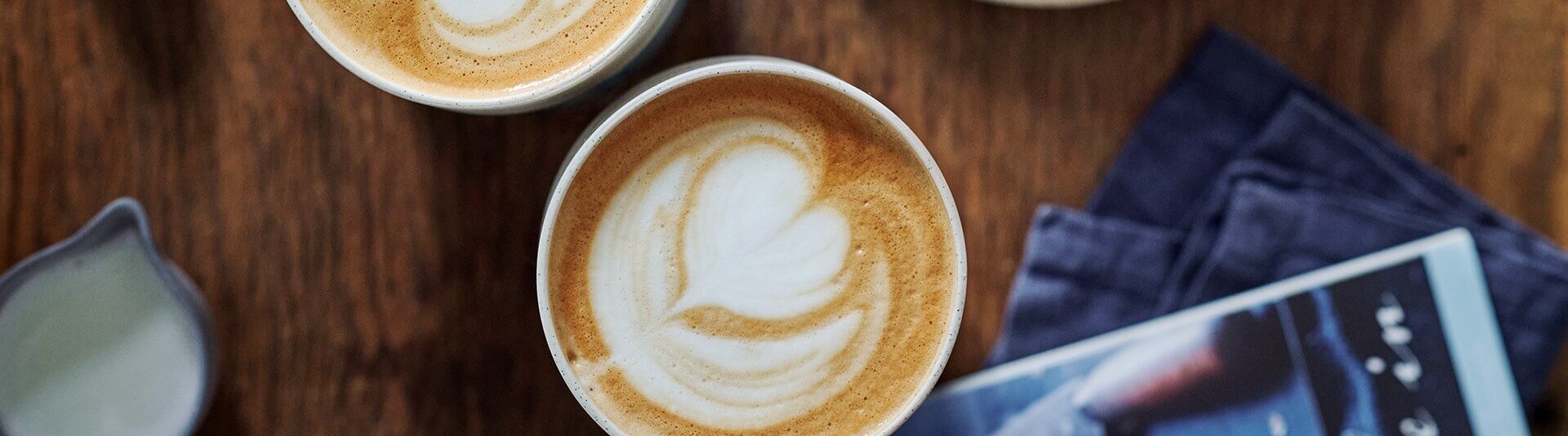 Zwei Tassen Milchkaffee auf einem Holztisch, dazu ein Gebäck und eine Zeitschrift für eine gemütliche Café-Atmosphäre.